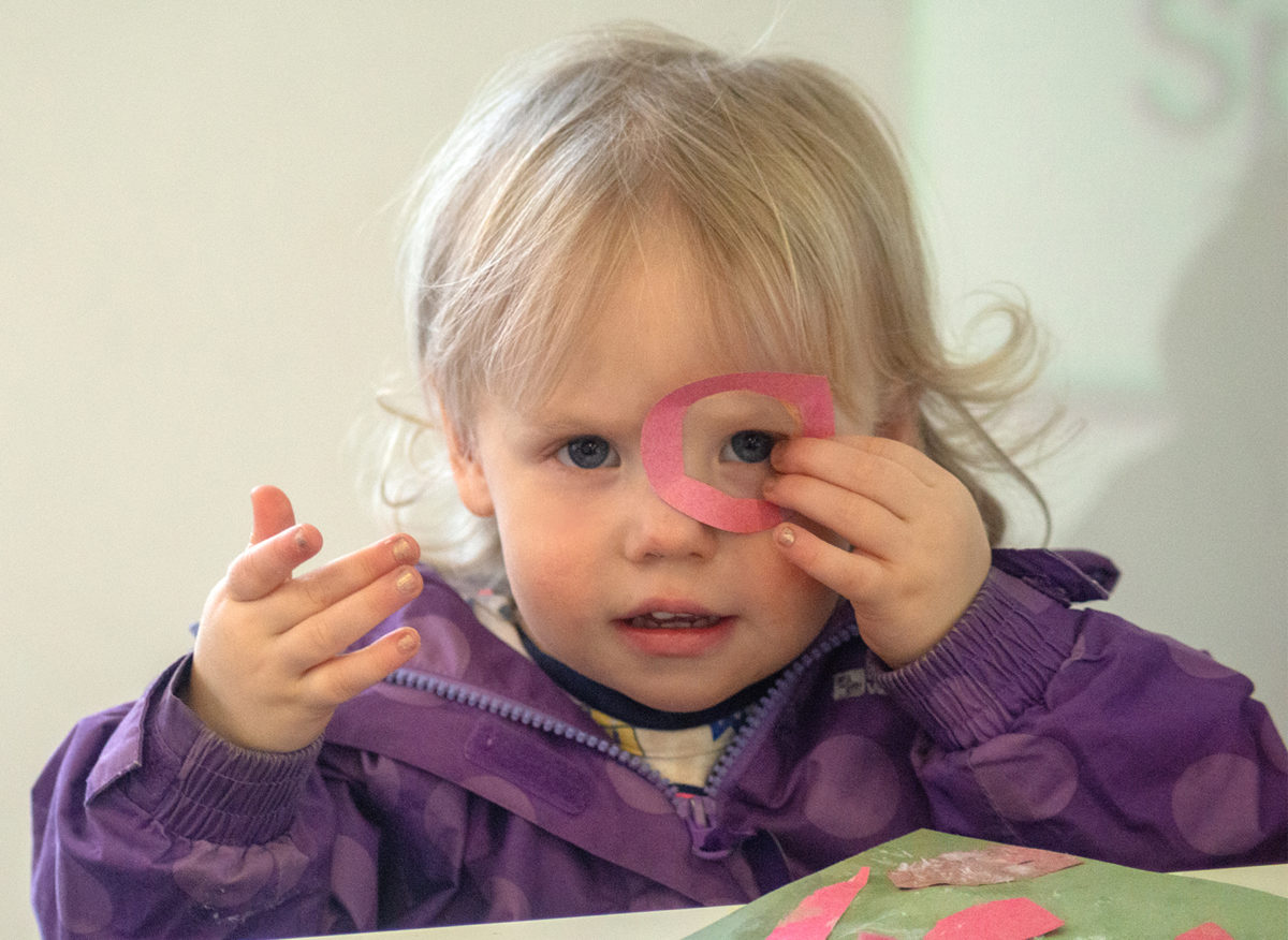 A toddler wearing a purple jacket peeks through a pink paper hoop she holds over her eye.
