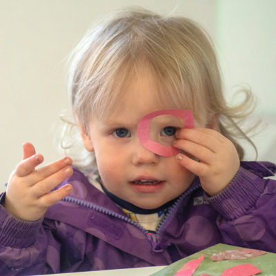 A toddler wearing a purple jacket peeks through a pink paper hoop she holds over her eye.