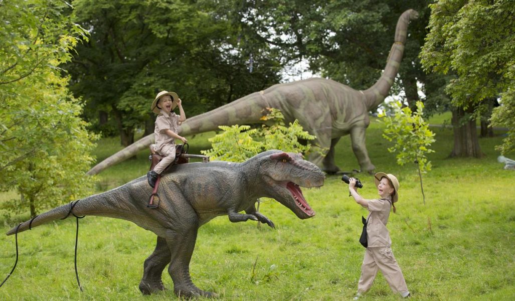 Two children dressed as explorers enjoy playing with a large model dinosaur.