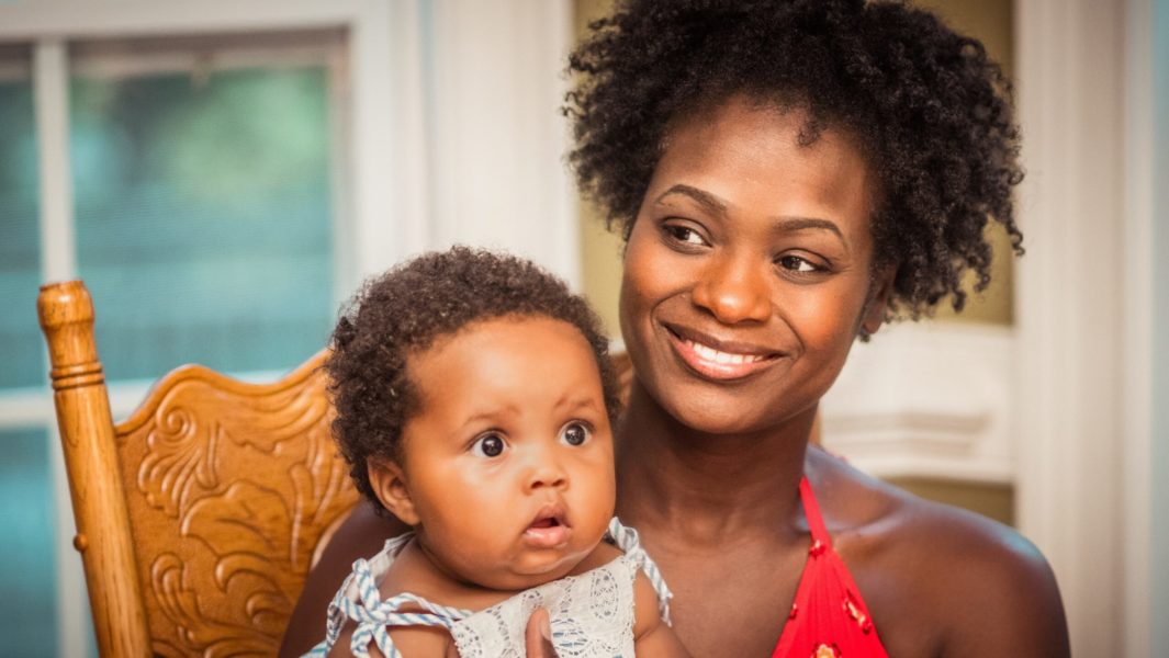 A lady smiles and sits with a baby on a chair.