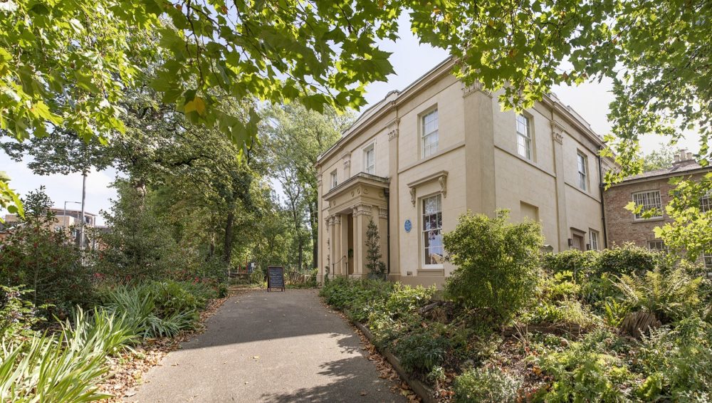 The building of Elizabeth Gaskell's House on a sunny day surrounded by trees.