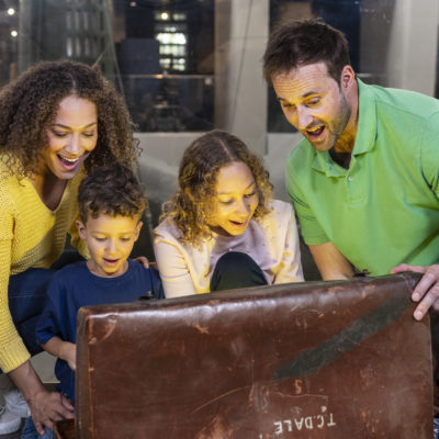 A family of four open an old looking brown leather chest and a light shines out of it. They look excited and happy.