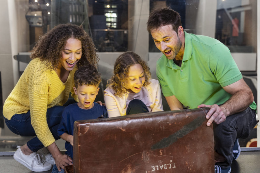 A family of four open an old looking brown leather chest and a light shines out of it. They look excited and happy.