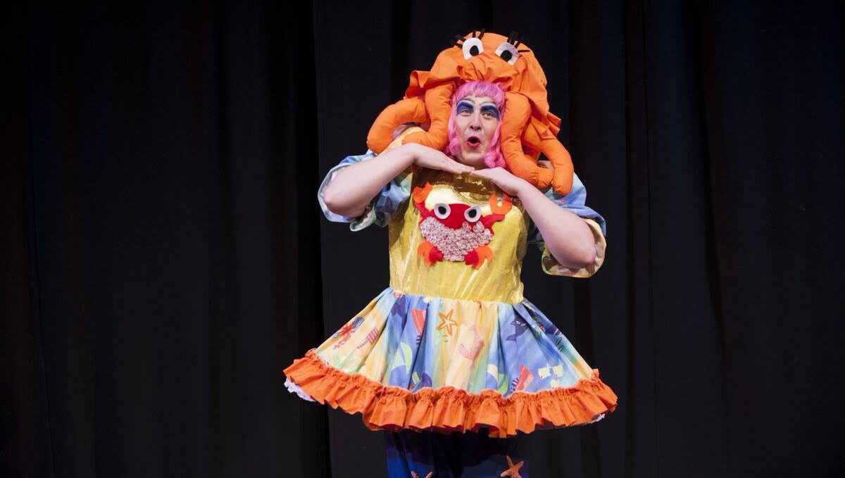 A pantomime dame - dressed in a yellow and blue dress with an orange octopus wig -stands with their hands underneath their chin.