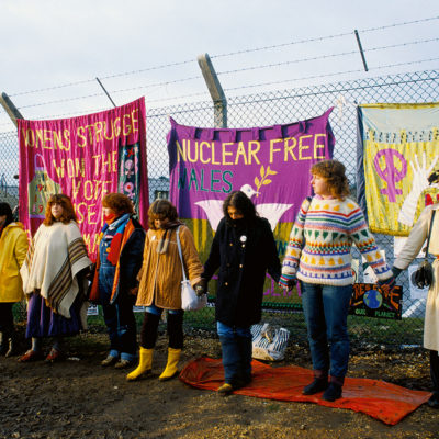 Womens Peace Camp protestors holding hands encircling USAF Base perimeter fence where three banners hang behind
