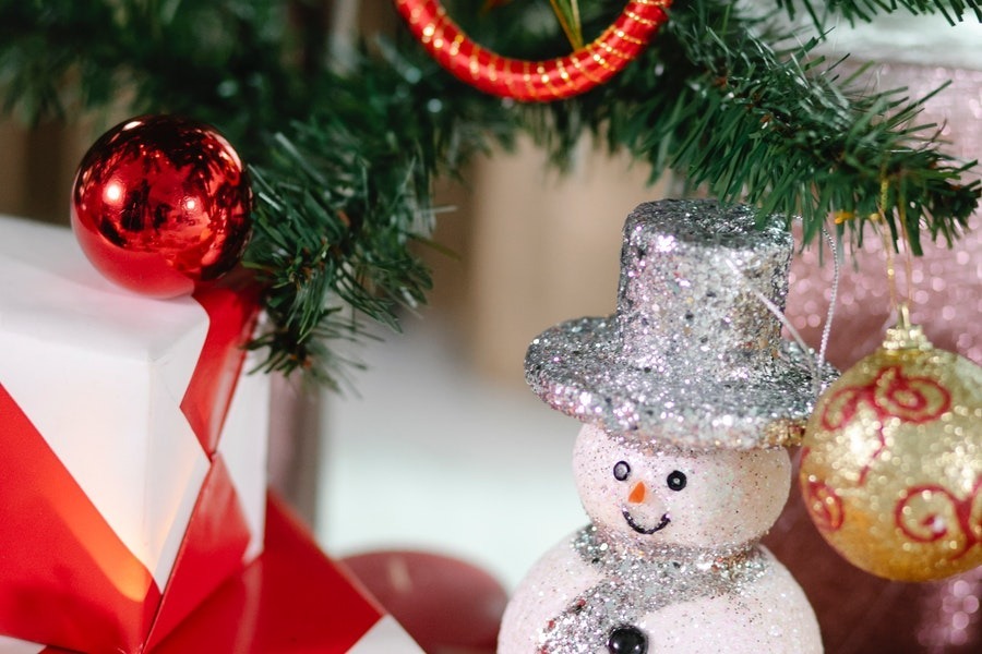 A sparkly snowman ornament with a silver hat and scarf sits beneath the branch of a Christmas tree.