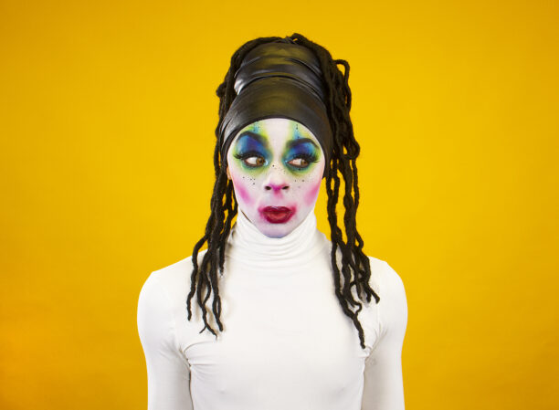 A person stands in front of a yellow background. They wear a white polar neck and a black hair band. They have long black hair and bright make-up on their face.