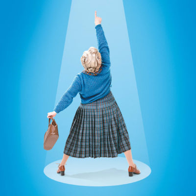 A woman wearing a green tartan skirt, a blue jumper and brown shoes holds a brown handbag and points to the sky.