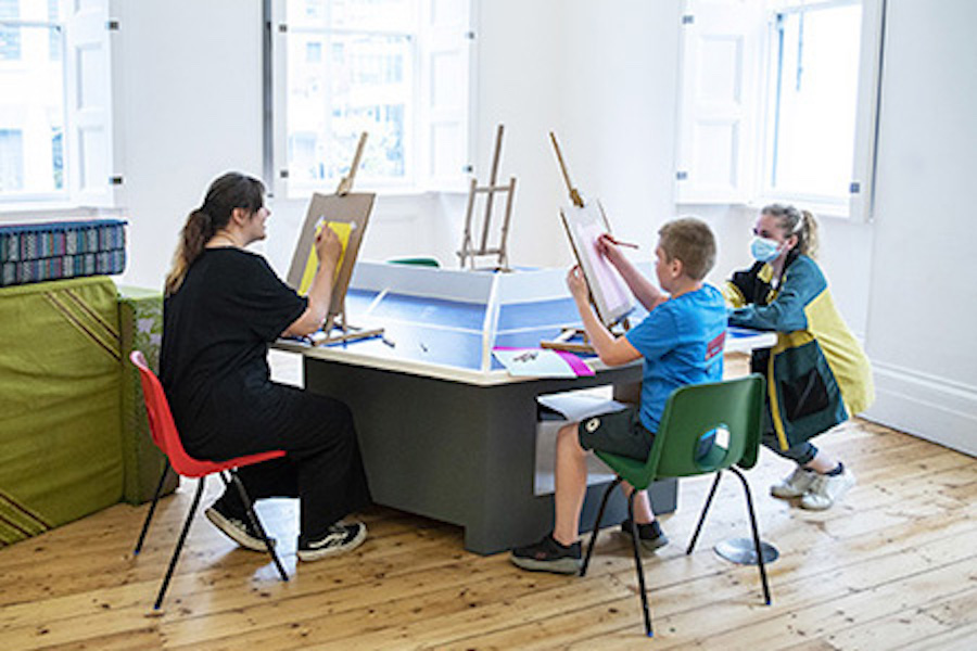 Three people sit at easels around a table making their own artwork.