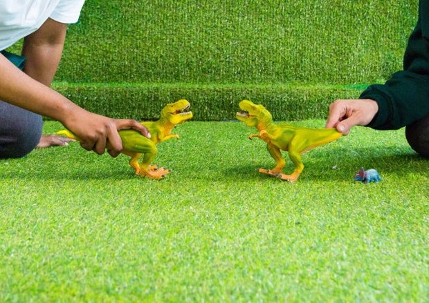 two green plastic toy dinosaurs stand on artificial grass - they are each held by a hand.