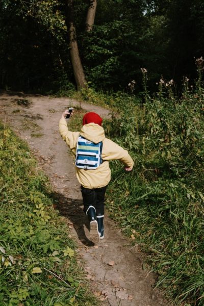 A small child wearing a yellow coat and a backpackruns down a pathway in a park