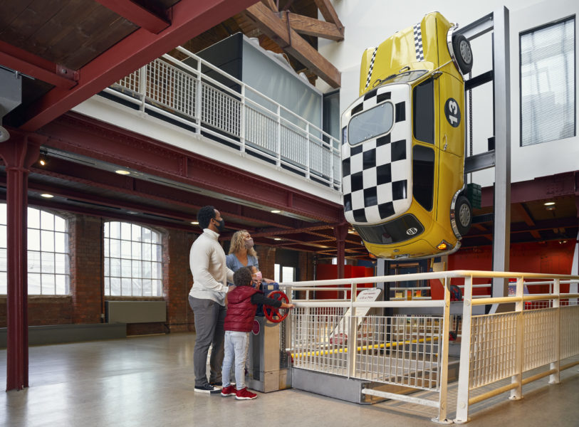 A family stand looking at an exhibition of a yellow car with a chequered roof.