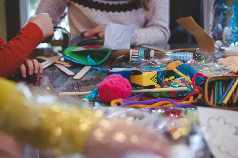 table top full of crafting materials, including crayons, pipe cleaners, coloured pencils, pompons, stickers, and lollypop sticks. The image also includes a couple of children crafting. You can only see part of the childrens torsos, arms and hand.