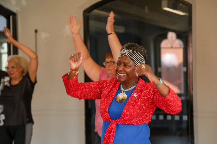 A smiling, Black, older woman wearing a red and blue top lifts her arms above her head in a dance