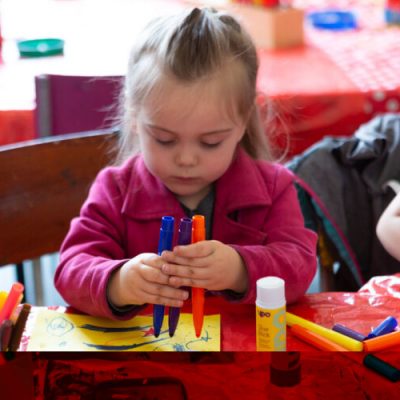 A small child holds three coloured felt tip pens on a piece of yellow paper.