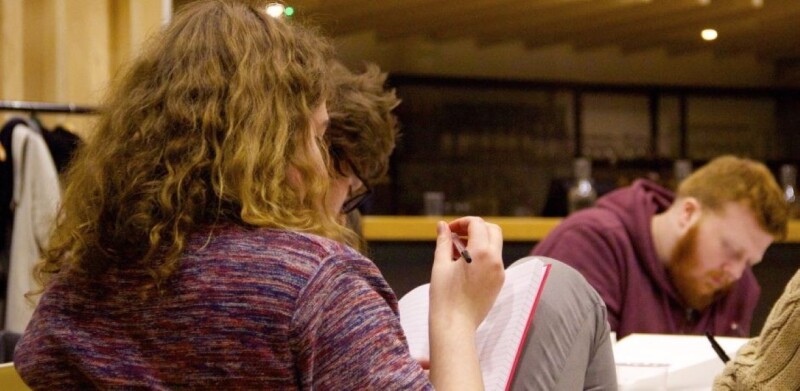 A young person with wavy brown hair sits with a pen and notepad writing