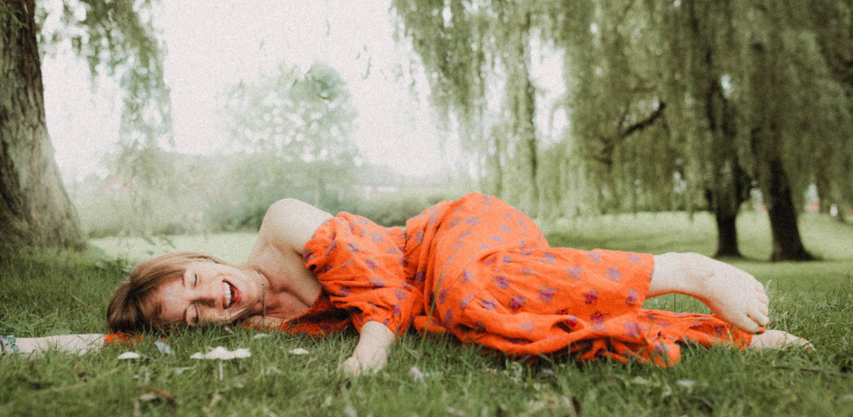 An adult wearing a long orange dress lies down in a grassy area.