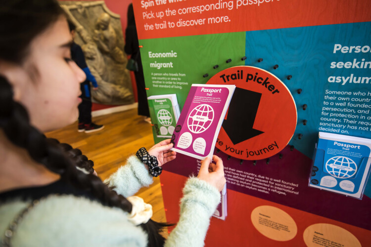 Child holding a Passport Trail activity booklet next to the trail picker wheel on the Migration a human story installation information board at People's History Museum